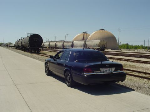 1999 Ford Crown Victoria - A black sedan is parked on a paved surface next to a train track with multiple rail cars and large domed structures in the background.