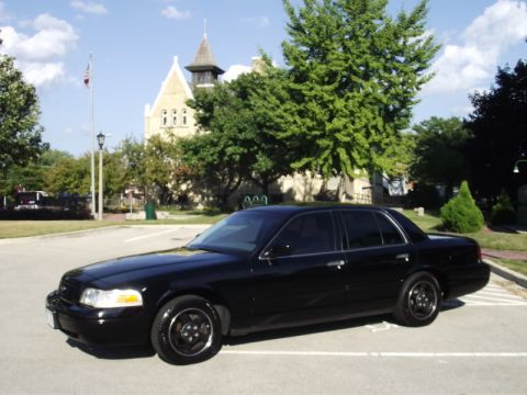 1999 Ford Crown Victoria - A black sedan is parked in a lot with a building and trees in the background. The car has aftermarket wheels.