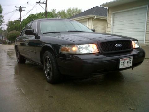 1999 Ford Crown Victoria - A black sedan sits on a wet road in front of a garage, its headlights illuminated.
