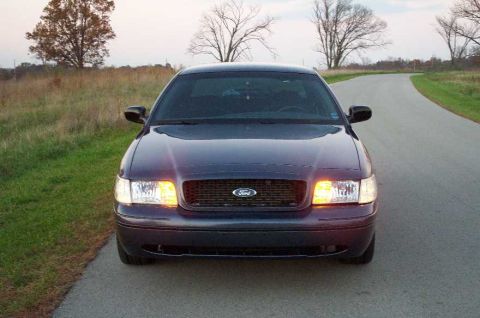 1998 Ford Crown Victoria - A dark plum/purple Ford Crown Victoria sedan photographed head-on with headlights illuminated, parked on a rural paved road with dry autumn…