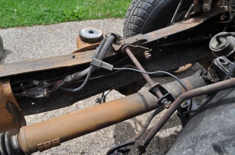 1986 Ford Bronco - Close-up view of the rusty frame and suspension components of a vehicle, with visible wiring and a tire in the background.