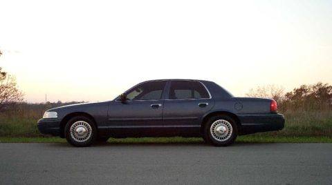 1998 Ford Crown Victoria - A dark blue Ford Crown Victoria full-size sedan shown in profile (driver's side), parked on pavement with dry grass and trees under a hazy…
