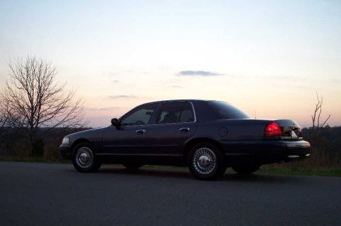 1998 Ford Crown Victoria - A dark blue/gray four-door Ford Crown Victoria sedan photographed from the rear three-quarter side angle at dusk, parked on a paved surface…