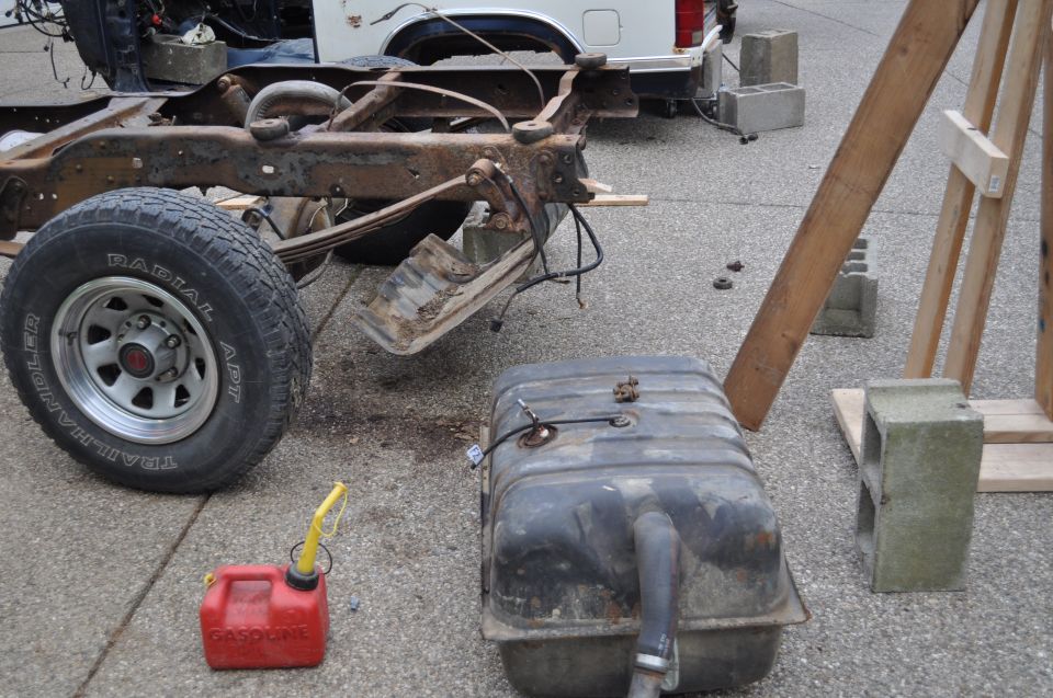 Rusted vehicle frame and leaf springs with a detached fuel tank and red gas can on asphalt