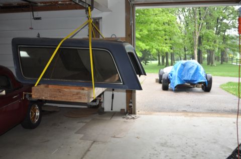 1986 Ford Bronco - A dark blue camper shell is suspended by yellow straps, with a red car visible below and another vehicle covered in a blue tarp in the back…