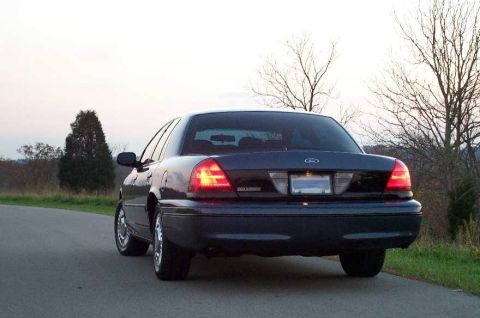 1998 Ford Crown Victoria - Rear view of a dark blue Ford Crown Victoria sedan parked on a paved rural road at dusk, with the driver-side taillight illuminated red and…