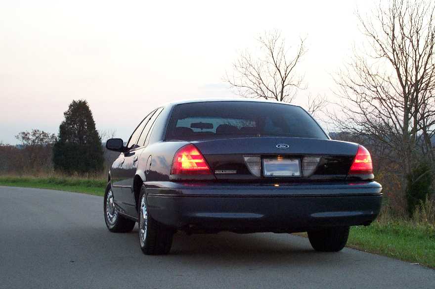 Rear of dark blue Ford Crown Victoria sedan on rural road at dusk, one taillight lit