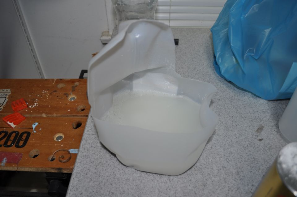 Plastic jug with white liquid on a counter next to a blue bag and a wooden surface with numbers