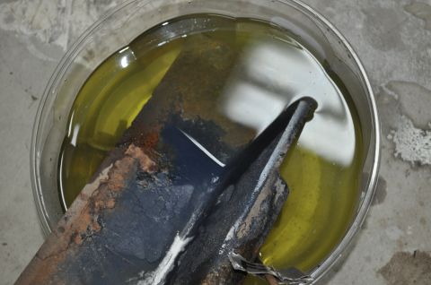 1986 Ford Bronco - A heavily rusted metal component is submerged in a yellow liquid, likely for rust removal, with a website logo in the corner.