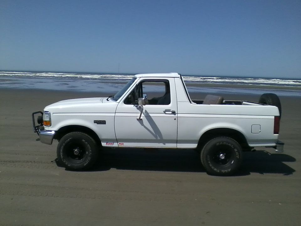 White Ford Bronco with removed rear top, black wheels and brush guard, parked on a beach