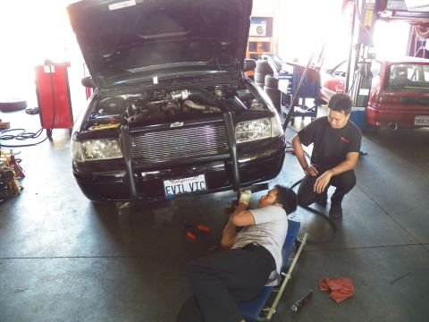 2003 Ford Crown Victoria - EVIL VIC Ford sedan in a garage bay, one tech on a creeper and another kneeling beside it