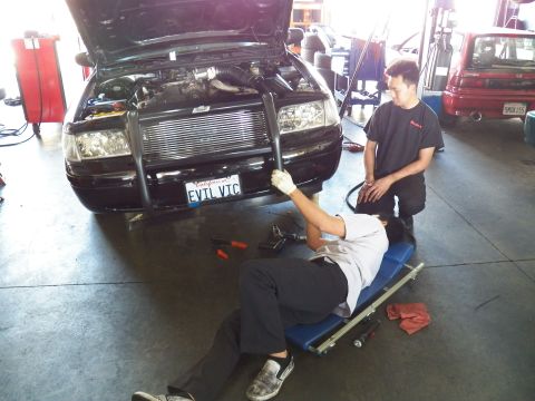 2003 Ford Crown Victoria - EVIL VIC Ford sedan with hood up, worker lying on a creeper reaching upward, tech standing by