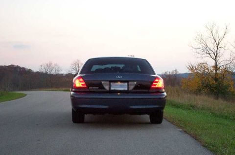 1998 Ford Crown Victoria - Rear view of a dark blue Ford Crown Victoria full-size sedan with illuminated red taillights, parked on a paved rural road at dusk with tre…