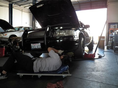 2003 Ford Crown Victoria - Worker slides under the lifted EVIL VIC Crown Victoria front end as light pours in from the bay door