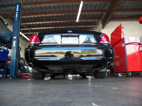 2003 Ford Crown Victoria - Ground-level shot of a black sedan's rear showing dual exhaust tips, chrome bumper and trunk spoiler.