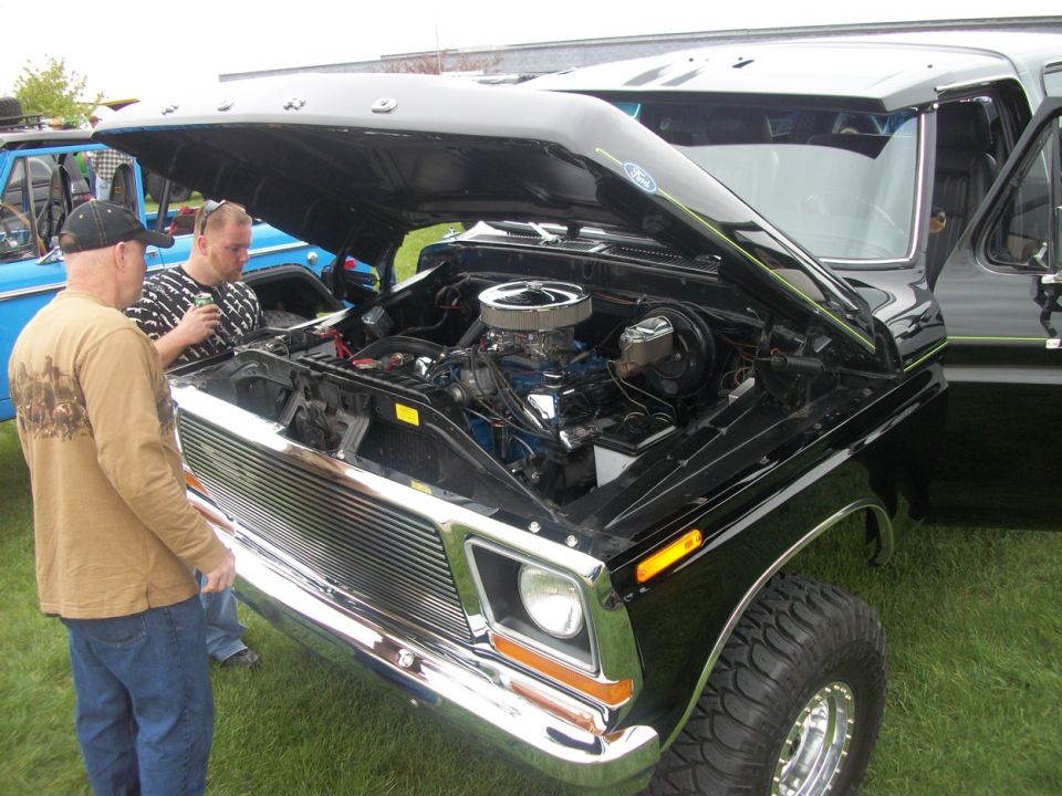 Two guys looking at a black Ford truck with hood open, showing the engine. Another blue Ford truck is parked nearby