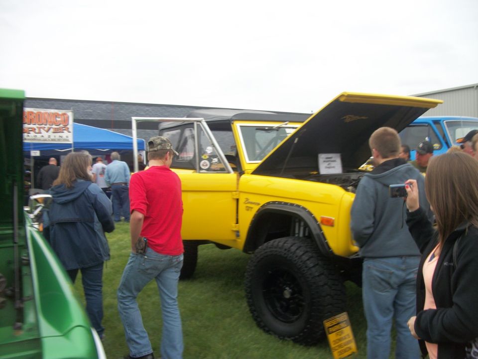 Yellow Ford Bronco with hood open, showing engine. "Bronco 1978" badge visible on fender. People looking at it