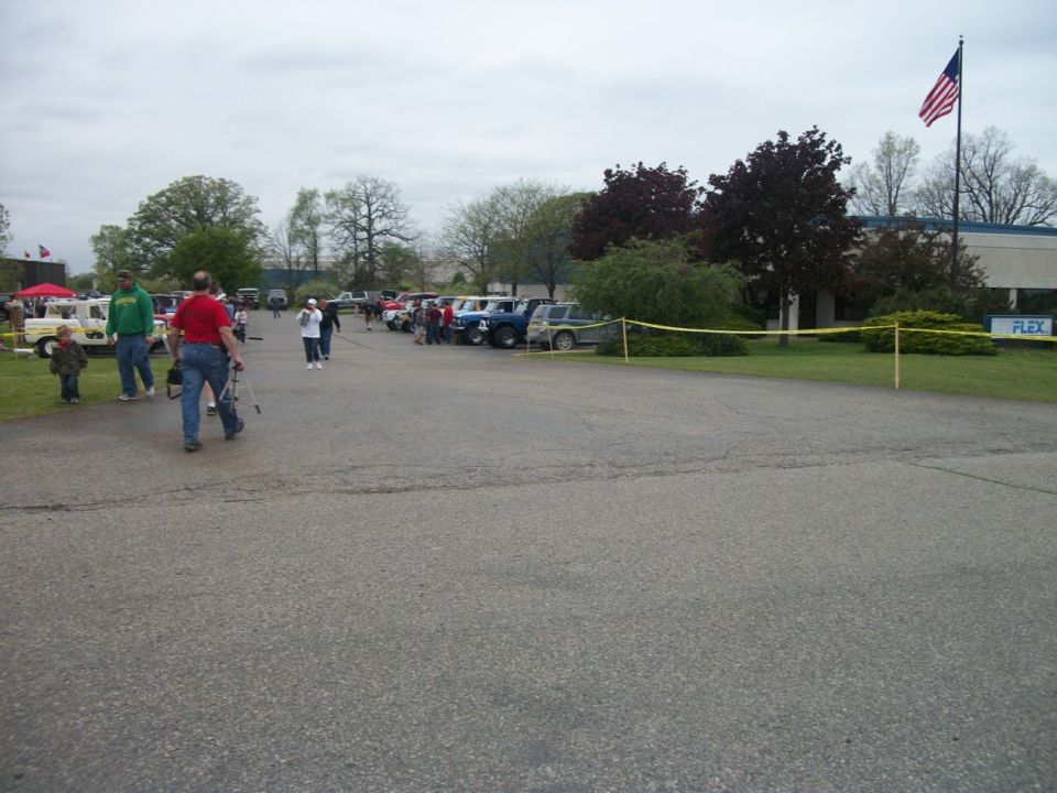 Outdoor event with people milling around parked cars and trucks, American flag flying