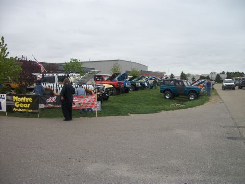 1996 Ford Bronco - A car show features multiple vehicles, including several Ford Broncos, displayed on a grassy area under an overcast sky.