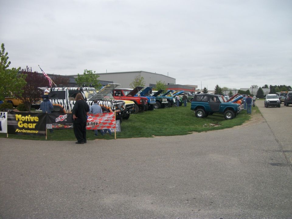 Several Broncos and other trucks lined up on grass with people around. One Bronco is teal, another has tiger stripes