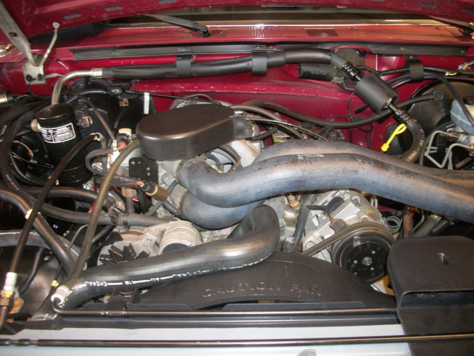 Engine bay with hoses, belts, and fan visible. Text on fan shroud reads 'CAUTION: FAN'