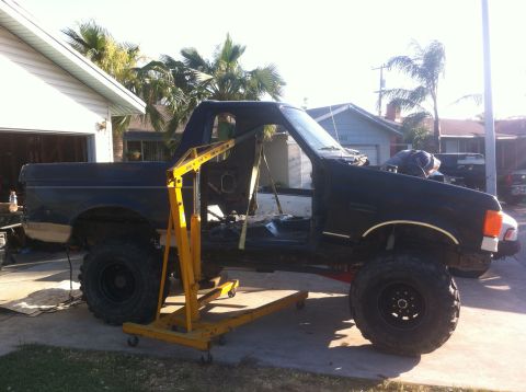 1989 Ford Bronco - A black Ford Bronco is partially disassembled in a driveway, with a yellow engine hoist positioned to lift the engine.