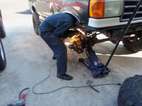 1968 Ford Bronco - A mechanic uses an angle grinder on a vehicle's front suspension while it is supported by a jack.