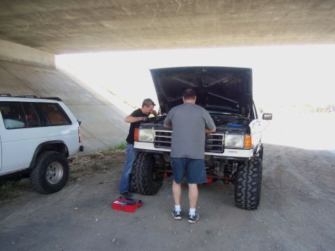 1968 Ford Bronco - Two men are working on the engine of a lifted SUV under a bridge, with another SUV parked nearby.