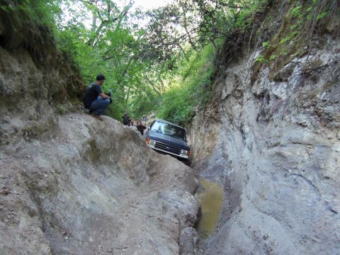 1968 Ford Bronco - A dark Ford truck with a chrome grille noses down into a narrow, deeply eroded off-road rut with muddy water pooled at the bottom, walls of…