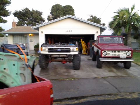 1968 Ford Bronco - A red and white 1968 Ford Bronco is parked in front of a garage, with a lifted white truck beside it.