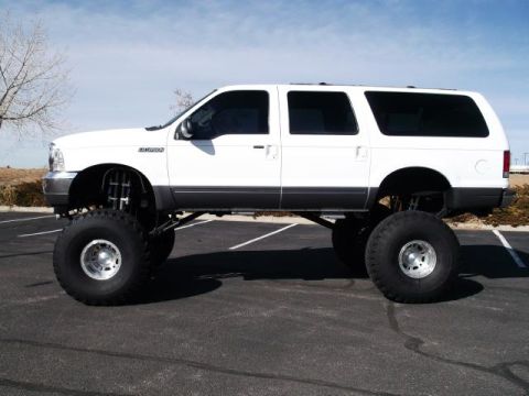 2001 Ford Excursion - A heavily modified white Ford Excursion SUV with oversized tires and a lifted suspension is parked on asphalt under a clear sky.