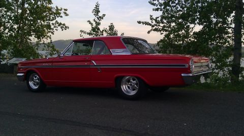 1964 Ford Fairlane - A red 1964 Ford Fairlane coupe is seen from its rear three-quarter angle, parked beside a road with trees and water in the background.