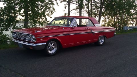 1964 Ford Fairlane - A red 1963 Ford Falcon sits parked on the side of a road next to a body of water.