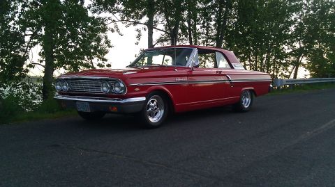 1964 Ford Fairlane - A bright red 1964 Ford Fairlane is parked on the side of a road with trees and water in the background.