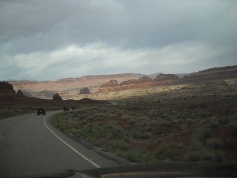 1986 Ford Bronco II - SUVs travel a road through a vast desert with rock formations under a dramatic sky.