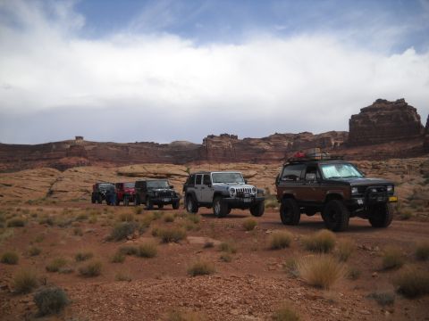 1986 Ford Bronco II - Several off-road vehicles, including a brown and black SUV and multiple Jeeps, travel through a desert landscape.