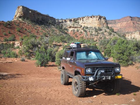 1986 Ford Bronco II - A brown 1986 Ford Bronco II is parked in a desert area with red rock formations in the background.