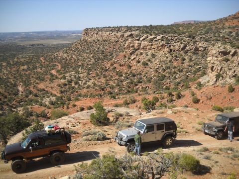 1986 Ford Bronco II - Three off-road vehicles navigate a dusty trail through a desert landscape with red rock cliffs.