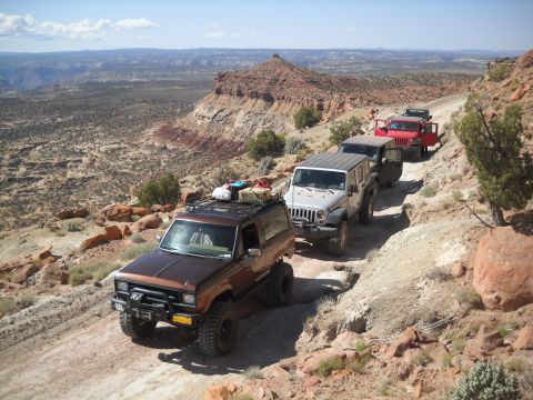 1986 Ford Bronco II - A convoy of off-road vehicles, including a 1986 Ford Bronco II, navigates a challenging dirt track through a desert canyon.
