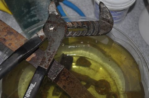 1986 Ford Bronco - Metal parts undergoing rust removal treatment in a clear container filled with a yellow solution.
