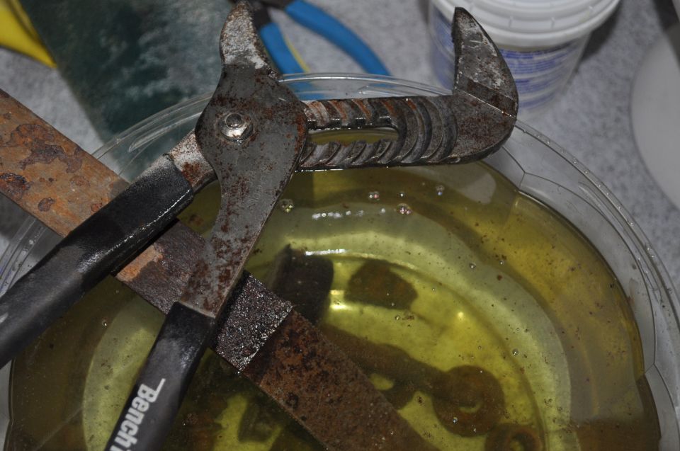 Rusted metal parts soaking in yellow liquid, with pliers and a chisel visible