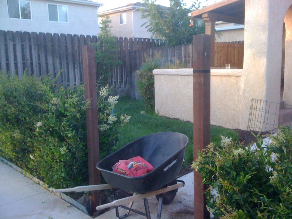 Wheelbarrow with two red bags of soil, on a paved surface next to a wooden fence and bushes