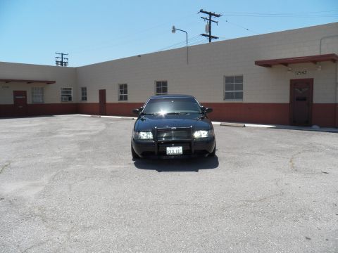 2003 Ford Crown Victoria - Distant front view of a black sedan parked alone in a wide industrial parking lot.