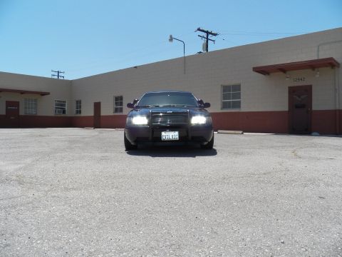 2003 Ford Crown Victoria - Black sedan photographed head-on with both headlights on against a beige warehouse.