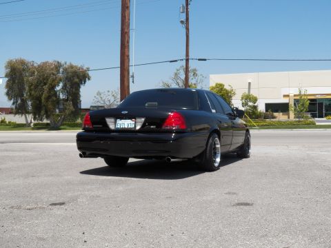 2003 Ford Crown Victoria - Black Crown Victoria shot from behind on a street, dual exhaust and chrome-lip wheels visible.
