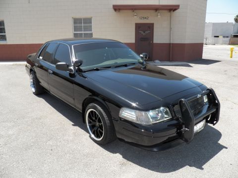 2003 Ford Crown Victoria - Black Crown Victoria with chrome push bar and pillar spotlight parked by a building marked 12942.