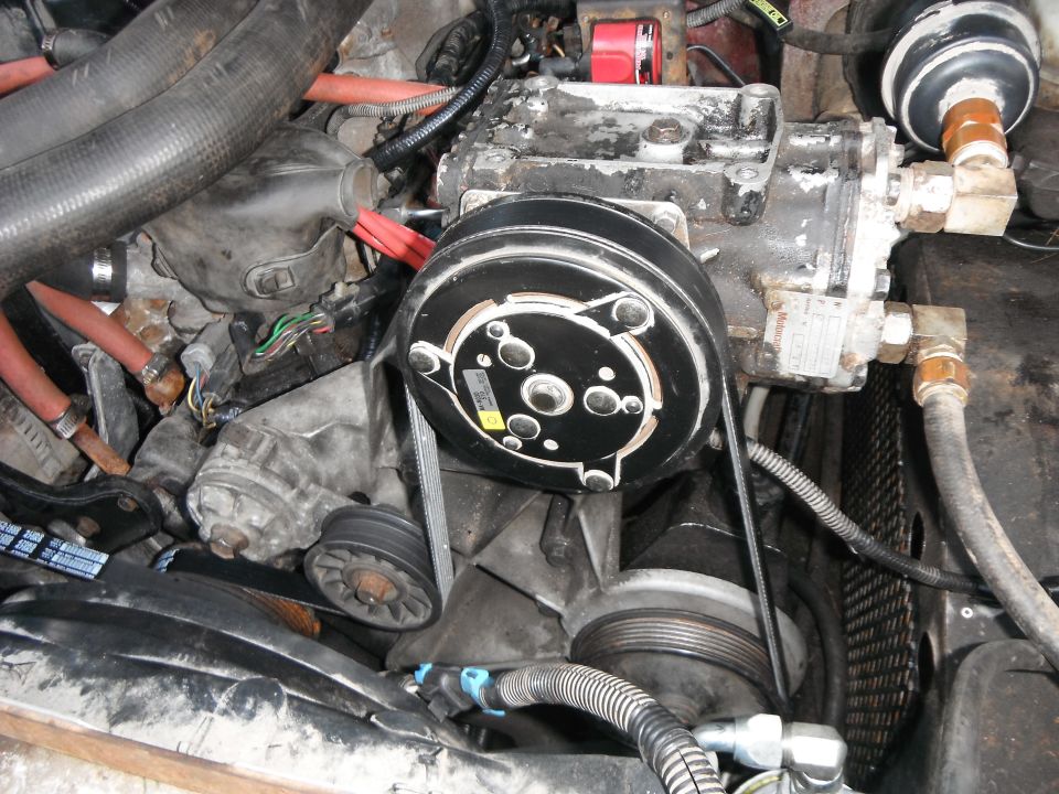 Close-up of engine bay showing AC compressor, belts, hoses, and pulleys. Supermotors.net logo visible