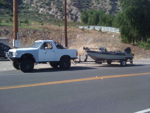1990 Ford Bronco - A modified pickup truck tows a Tracker boat on a trailer down a two-lane road.