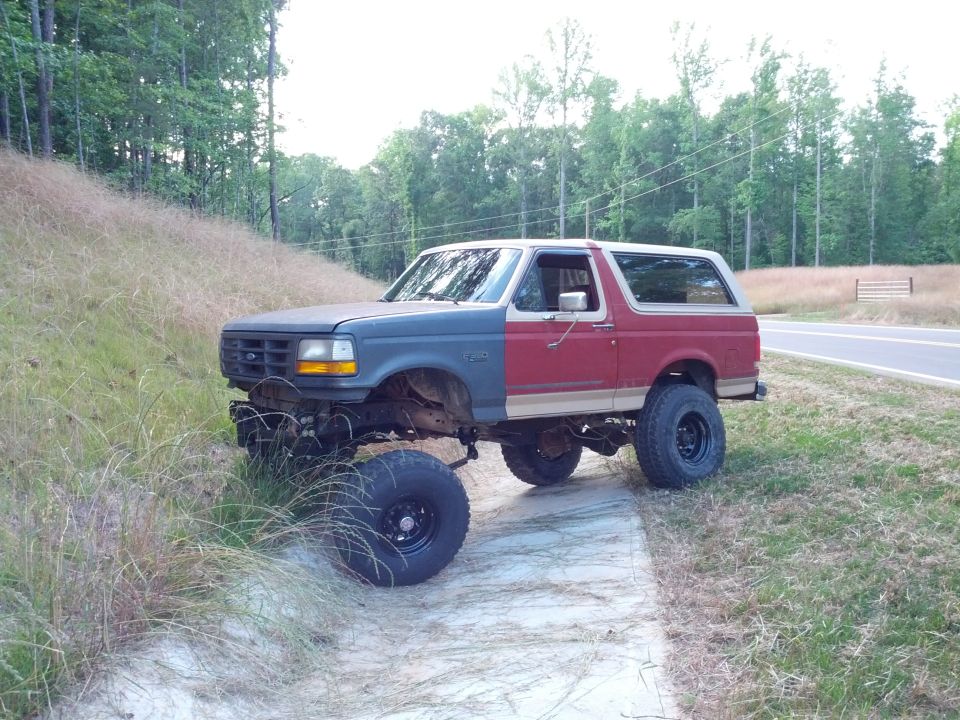Two-tone lifted Ford Bronco with mismatched front end, parked on a dirt path next to a road
