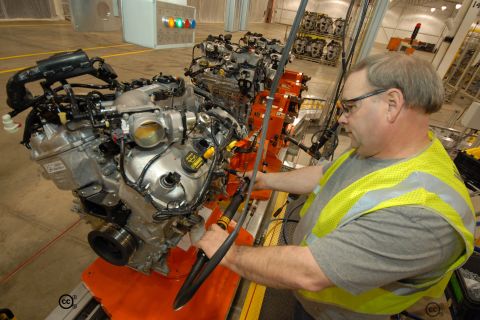 1997 Ford Taurus - A worker in a high-visibility vest works on a car engine on an assembly line. Multiple engines are visible in the background.